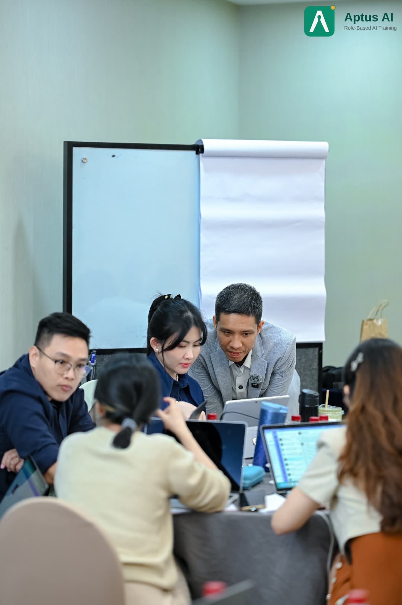 Giảng viên cúi xuống cùng một học viên xem bài thực hành trên laptop, các học viên khác làm việc quanh bàn